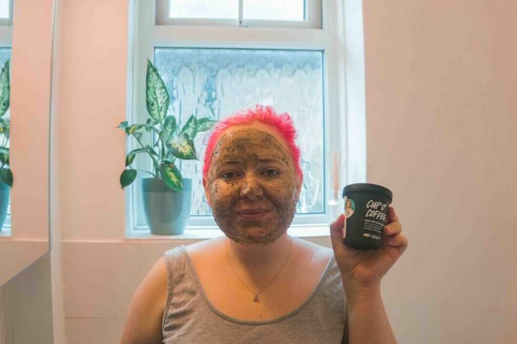 Kat with a coffee scrub on her face in the bathroom, holding a pot of cup of coffee from Lush. Behind her is a window and plant. 